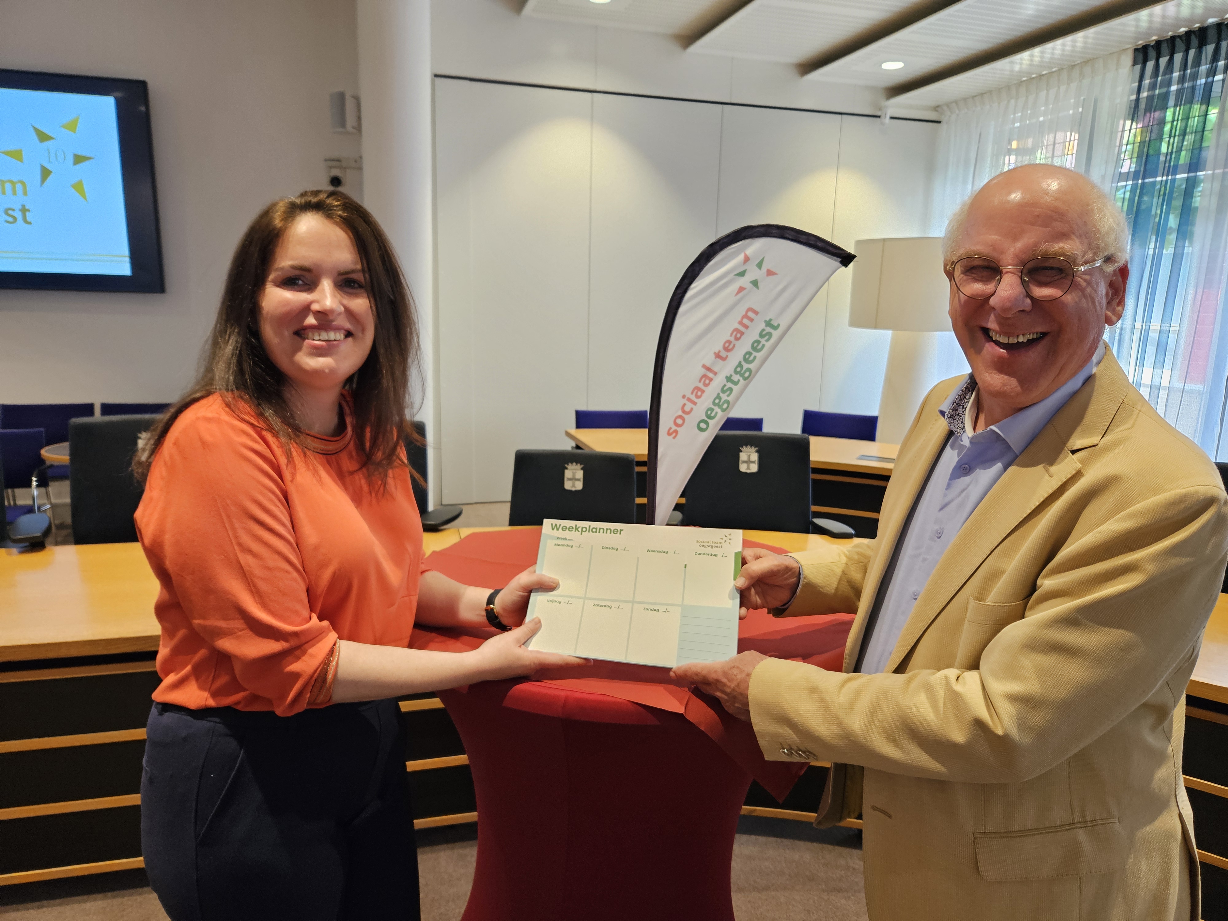 wethouder Tim van Tongeren en Astrid ter Horst, directeur van Radius schudden elkaar de hand en ze ontvangt de weekplanner, jubileumcadeautje