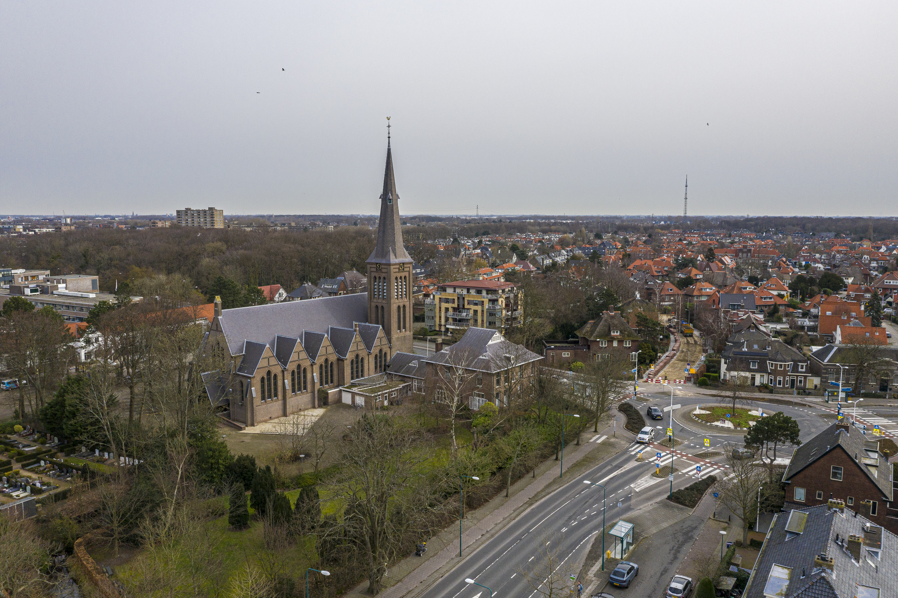 Luchtfoto van een grote bakstenen kerk met hoge toren, omringd door woonhuizen, bomen en een rotonde.