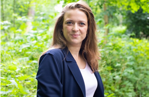 De persoon is een jonge vrouw met lang bruin haar en lichte ogen. Ze staat buiten, vermoedelijk in een bosrijke omgeving.