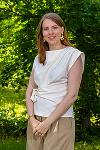 M. van Driel (Melanie), 2e locoburgemeester in witte blouse en bruine broek met groene struiken op de achtergrond.