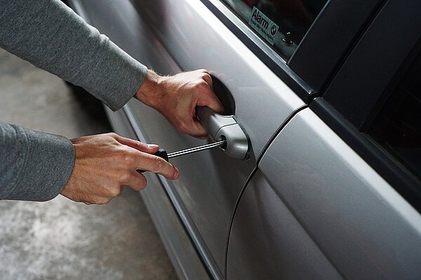 Foto van een grijze auto waar iemand met een schroevendraaier het deurslot probeert open te maken