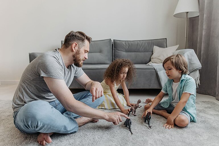 Afbeelding van een volwassen met twee kinderen die spelen met dinosaurussen in een huiskamer