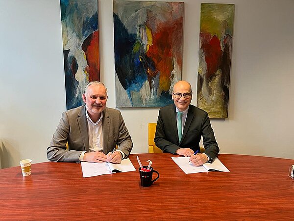 De heer van Pel, directeur M. en burgemeester Emile Jaensch zitten aan een oranje tafel voor het ondertekenen van het samenwerkingscontract